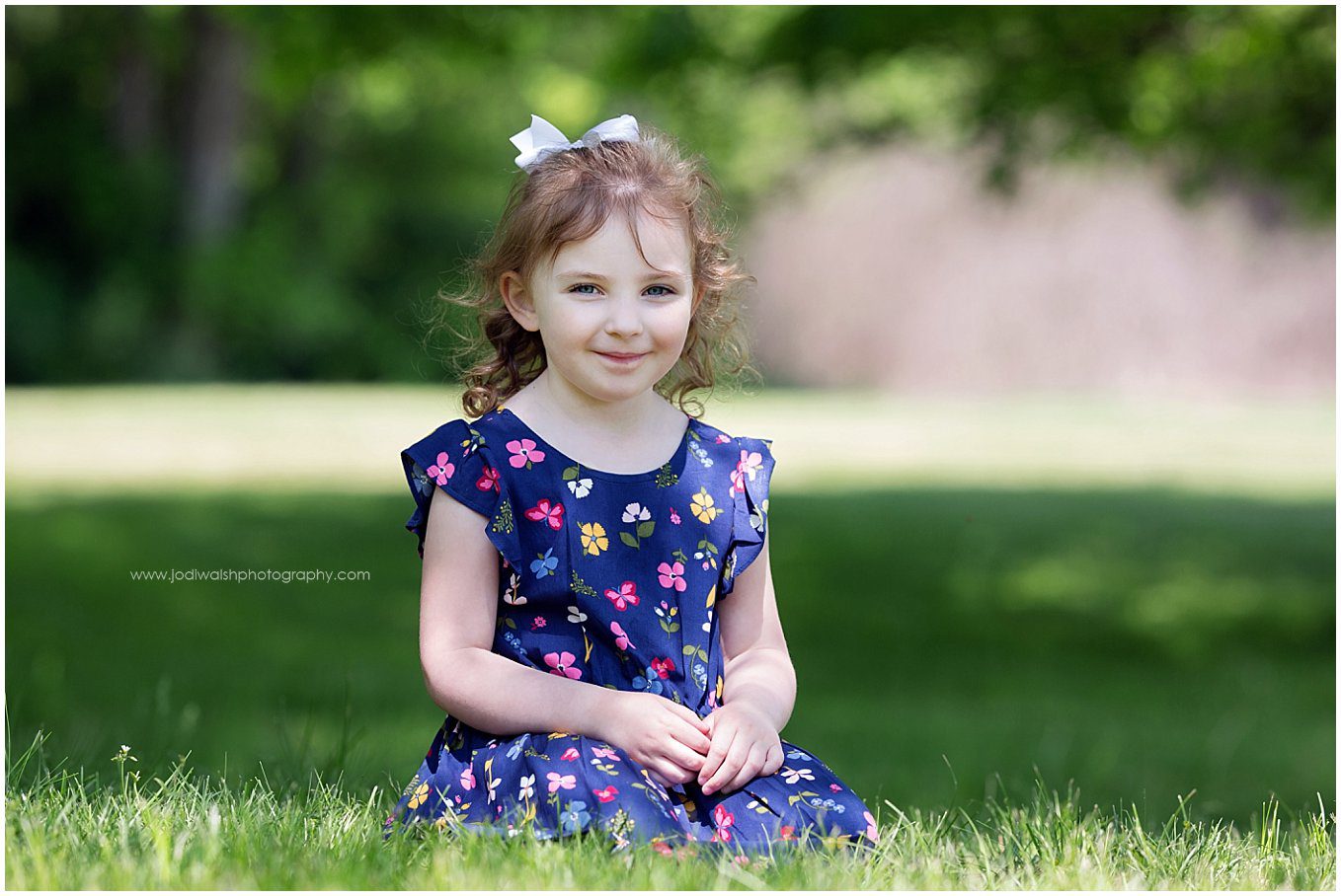 image of a little girl wearing a navy blue dress with flowers on it. She's sitting in the grass by a shade tree and has her hands in her lap. 