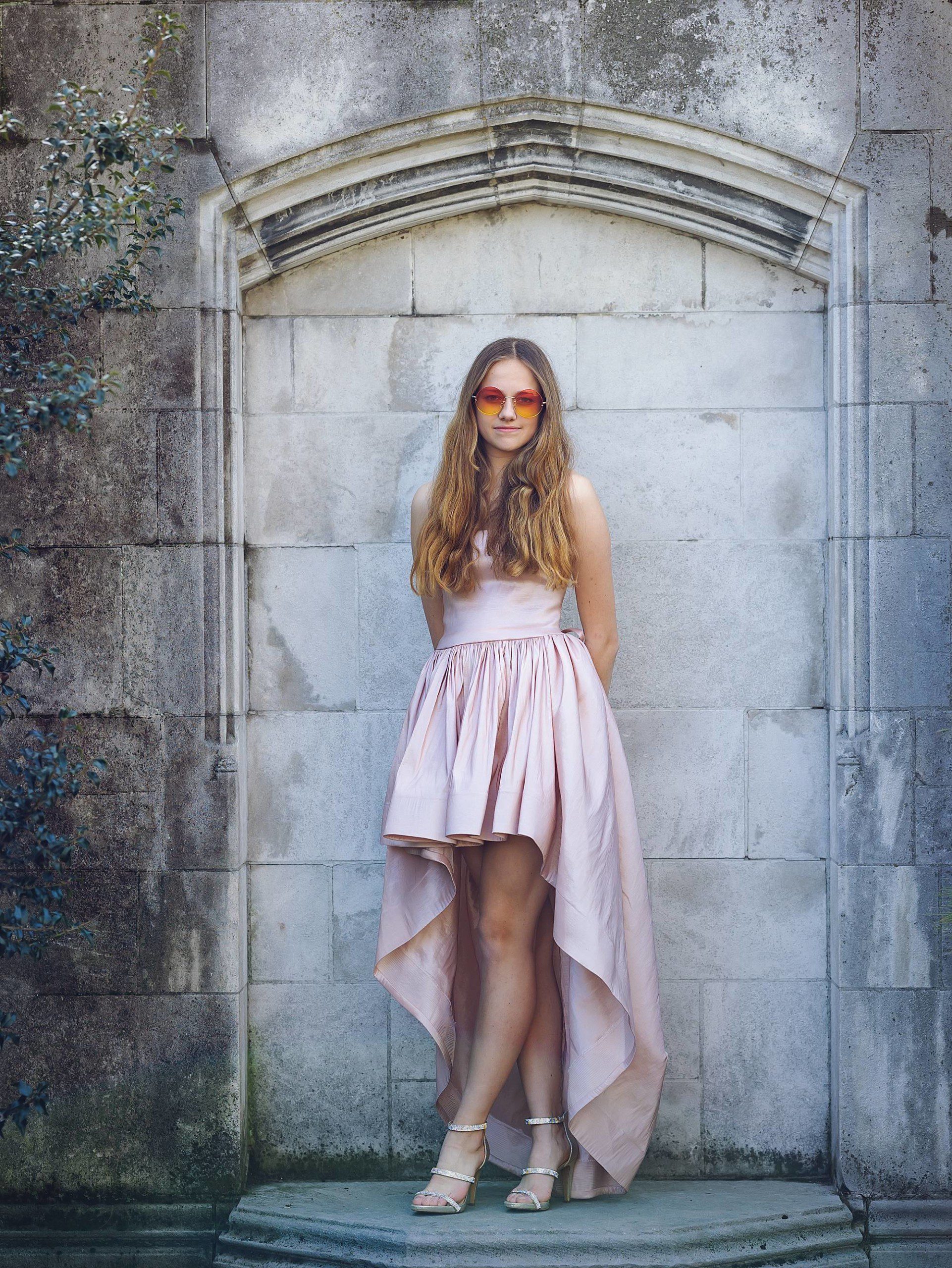 image of a senior girl in a high-low pink gown and rose sunglasses. She's standing on a low wall in a garden in Mellon Park, Pittsburgh.