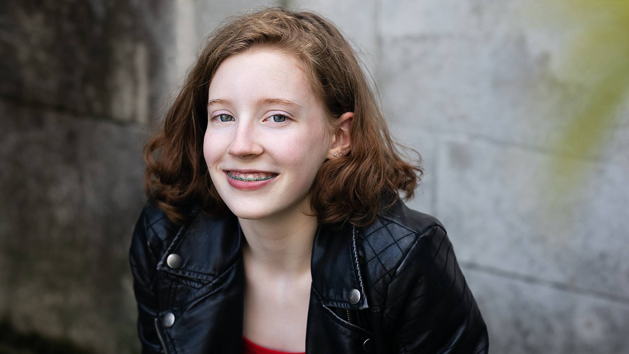 image of a teen with shoulder length wavy strawberry blonde hair. They are wearing a black leather jacket at Mellon Park in Pittsburgh.