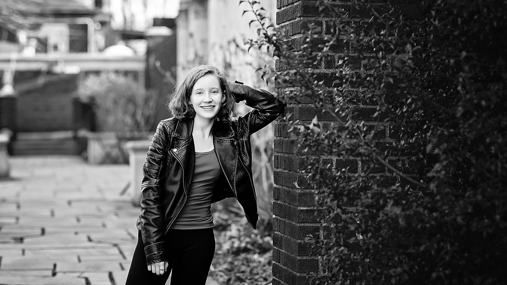 black and white image of a teen wearing a black leather jacket, leaning against a brick wall and smiling. They're in a garden at Mellon Park, Pittsburgh