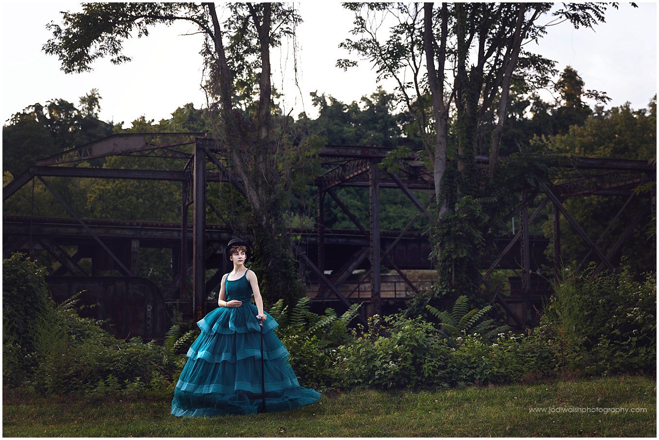 image of a teen wearing a teal gown with a black op hat.  They're standing in front of a rusted train trestle overgrown with trees and vines.