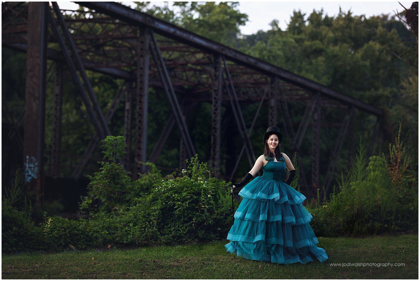 image of a teen girl wearing a teal gown with black gloves and a top hat.  She's standing in front of a rusted train trestle that's overgrown with trees and vines.