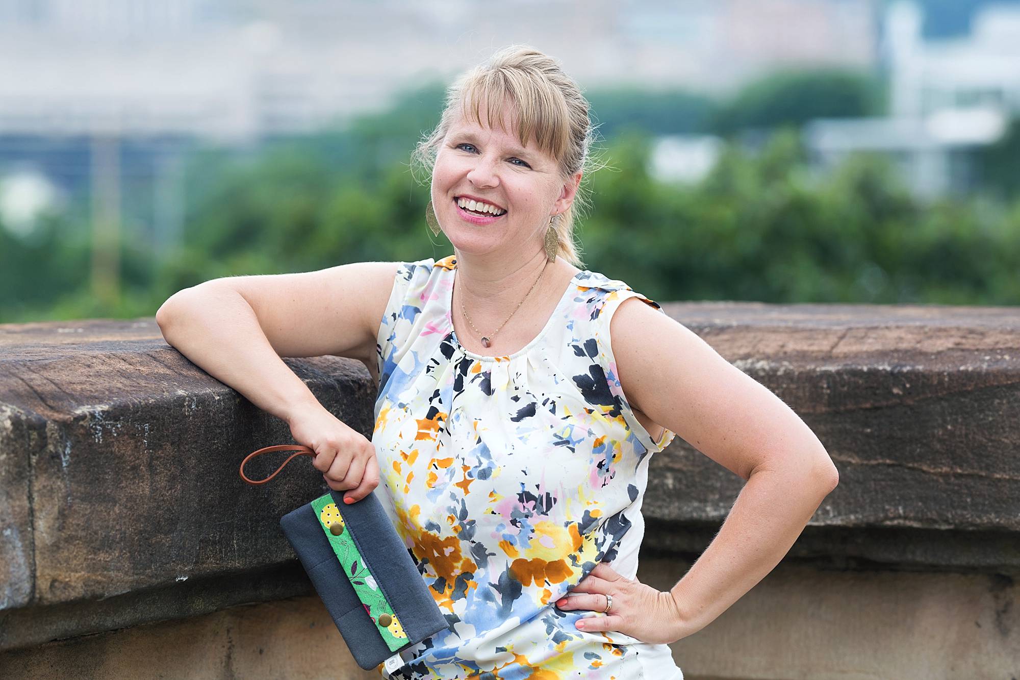 image of Theresa Grosh, designer behind Naptime Creations. She's standing on the Panther Hollow Bridge holding one of her custom designed wallets. She's a blond woman with her hair in a ponytail and is wearing a flowered, sleeveless shirt.