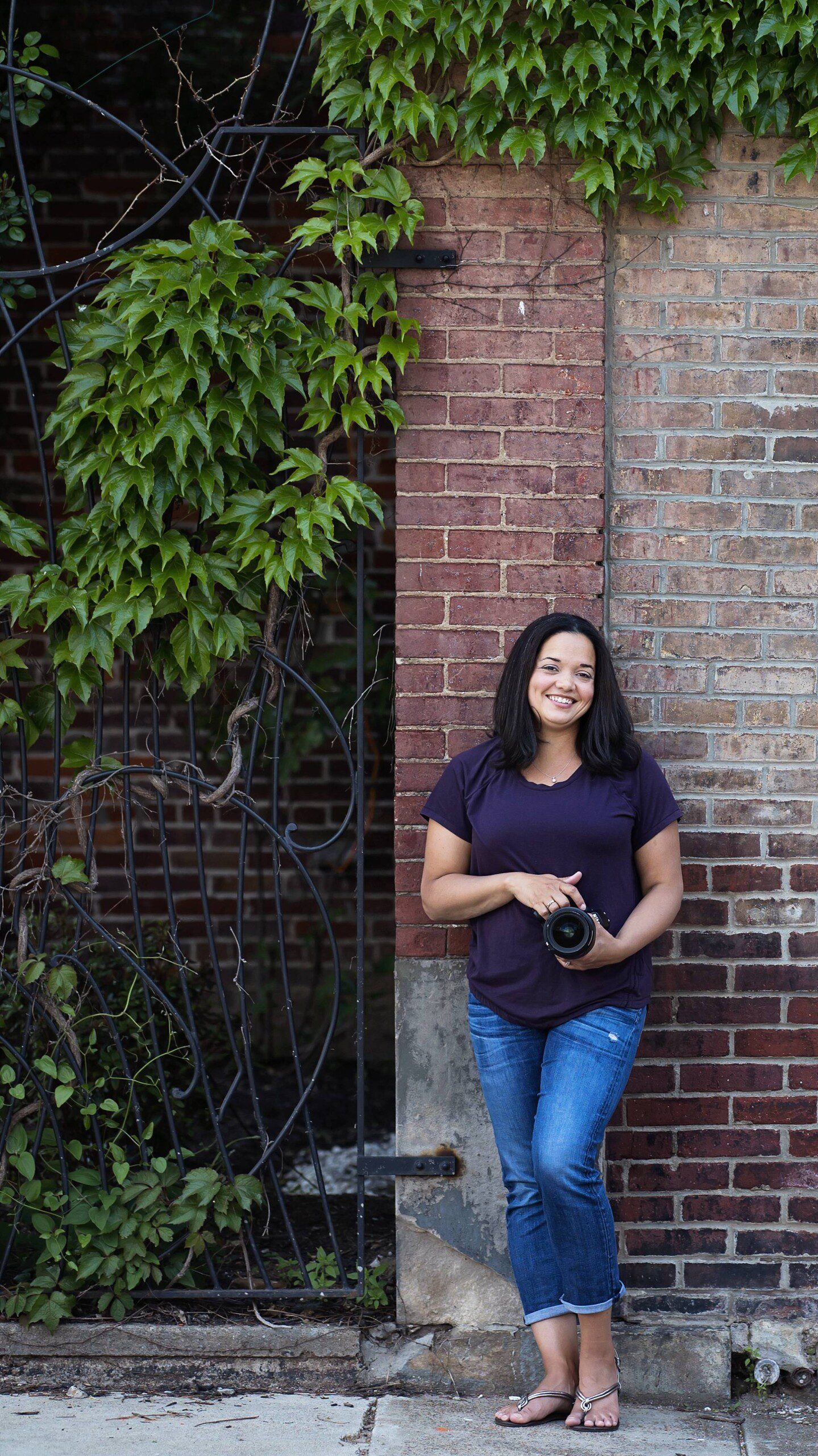 image of Rhaina Taylor, a Pittsburgh area portrait photographer. She's wearing jeans and a purple shirt as she leans against a brick wall in Lawrenceville