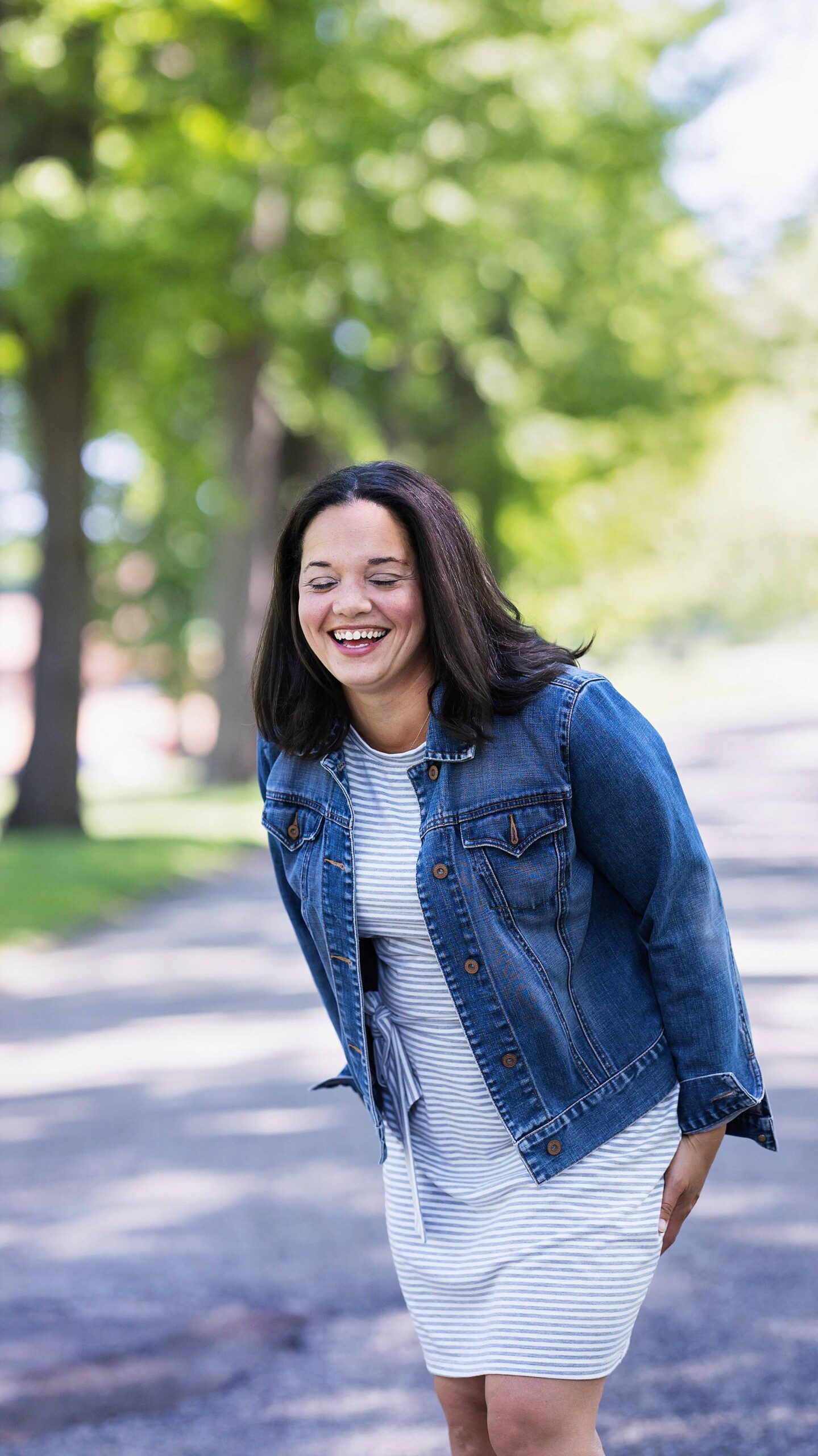 image of Pittsburgh area photographer, Rhaina Taylor. She's wearing a knit dress with a denim jacket and is laughing with her eyes closed while standing on a tree lined road.
