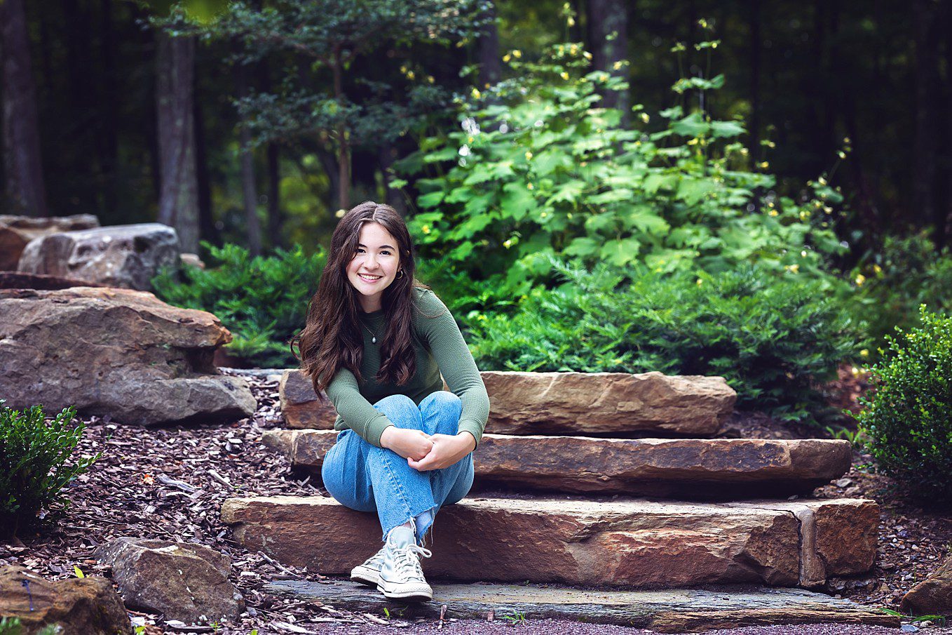 Senior girl wearing jeans and a green top, sitting on a set of stone steps in the woods around the Pittsburgh Botanic Gardens