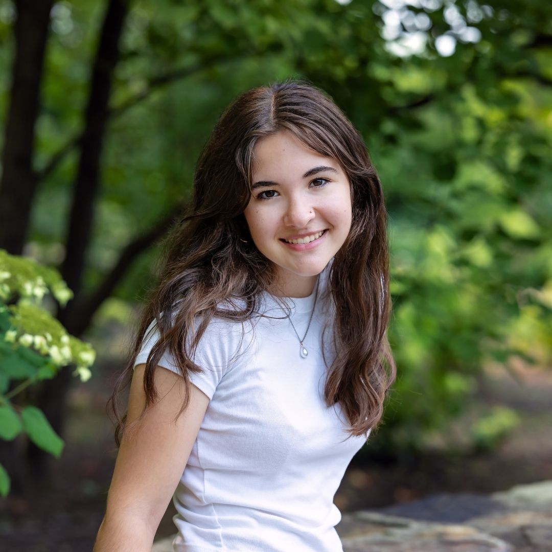 senior girl with long dark wavy hair, wearing a simple white t-shirt. she's leaning against a stone wall with in a wooded park at the Pittsburgh Botanic Gardens