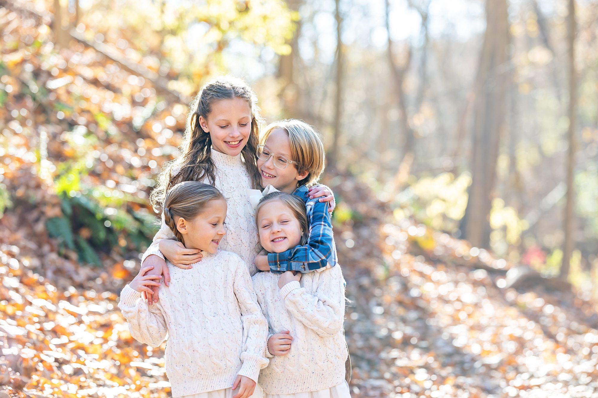 four siblings hugging while standing in the woods, one boy with two little sisters and one older sister. the girls are wearing white sweaters.