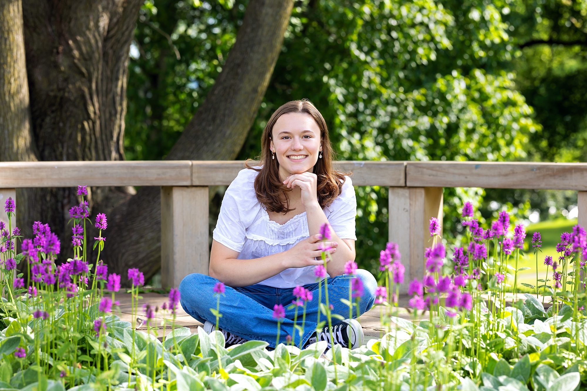 image of a senior girl wearing a white blouse and bluejeans, sitting cross legged surrounded by purple flowers in one of the gardens at Mellon Park
