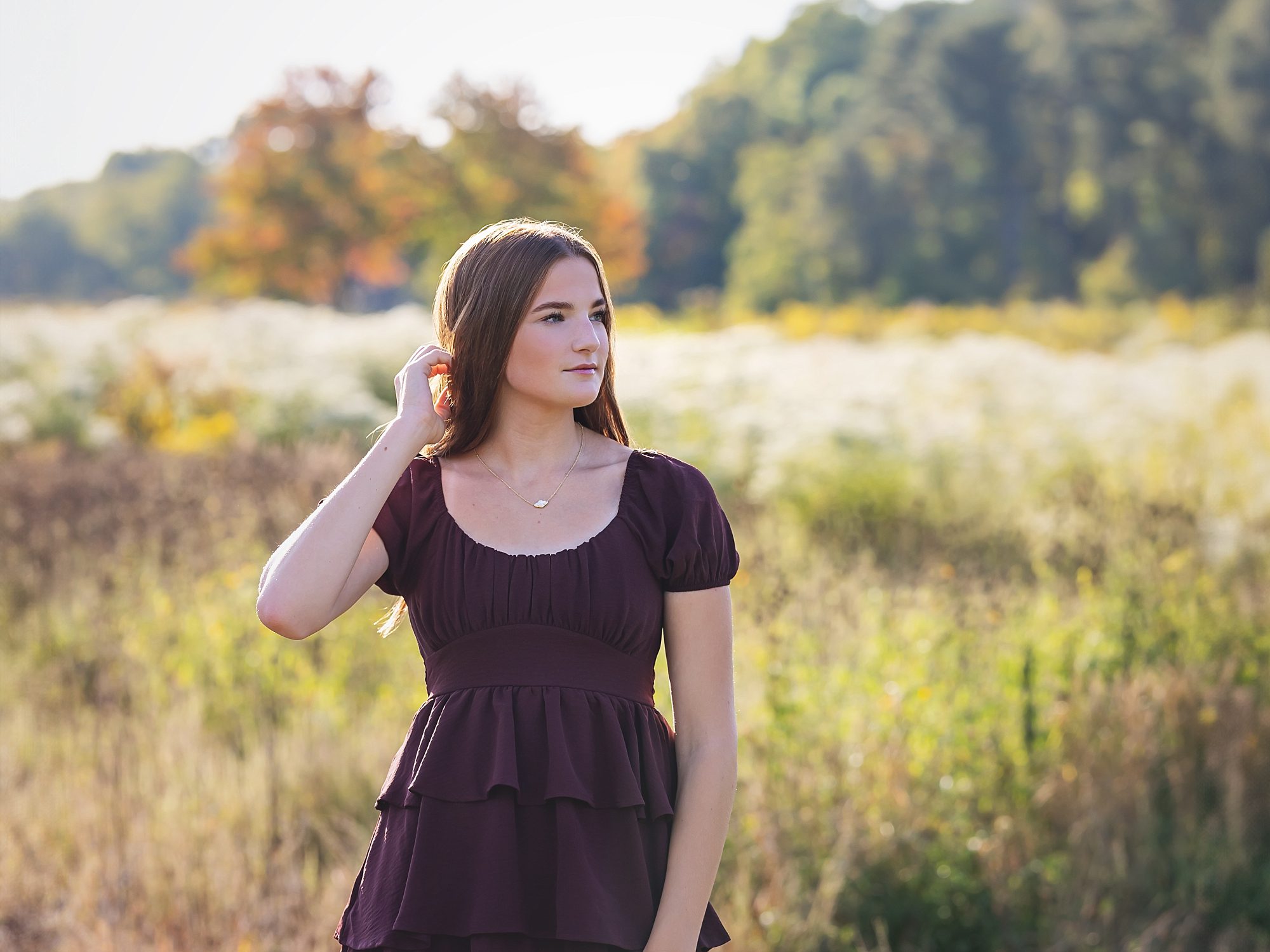 image of a teen girl in a burgundy dress standing in a field of tall grass and wildflowers.  She is looking off to the right as she tucks her long auburn hair behind her ear